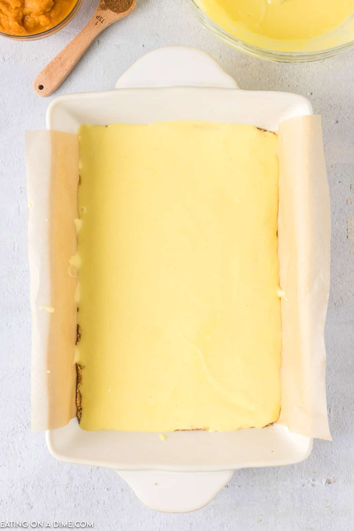 A white rectangular baking dish contains a smooth, evenly spread, creamy yellow layer of Pumpkin Cheesecake Bars. Parchment paper lines the dish. In the background are partially visible ingredients, including a wooden spoon and a bowl of yellow mixture on a light gray surface.