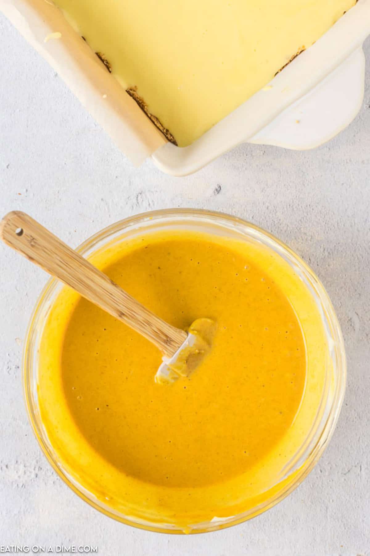 A glass bowl filled with smooth, yellow pumpkin pie mixture sits on a grey countertop. A wooden spatula rests inside the bowl. In the background, part of a baking dish with a baked crust is visible, ready to be filled with the mixture for delectable Pumpkin Cheesecake Bars.