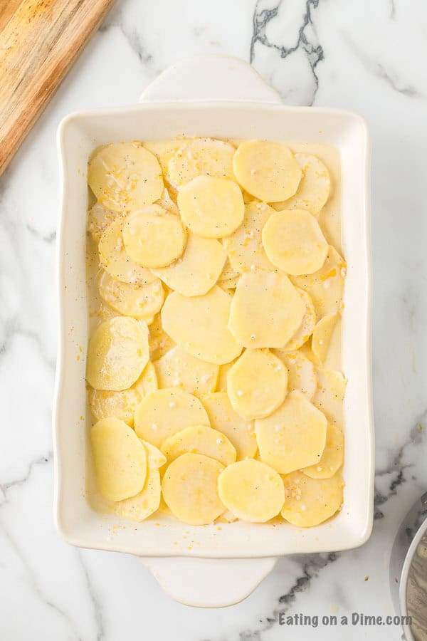 Overhead view of a white casserole dish filled with thinly sliced raw potatoes, lightly seasoned with salt and pepper on top. The dish is on a white marble countertop with part of a wooden cutting board visible in the corner. This sets the stage for a delightful Cheesy Scalloped Potatoes Recipe.
