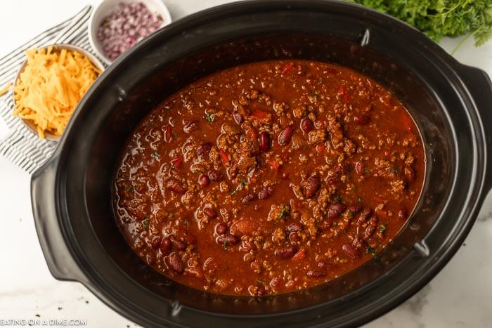 Cooked cowboy chili in a crock pot with shredded cheese and red onions next to it. 