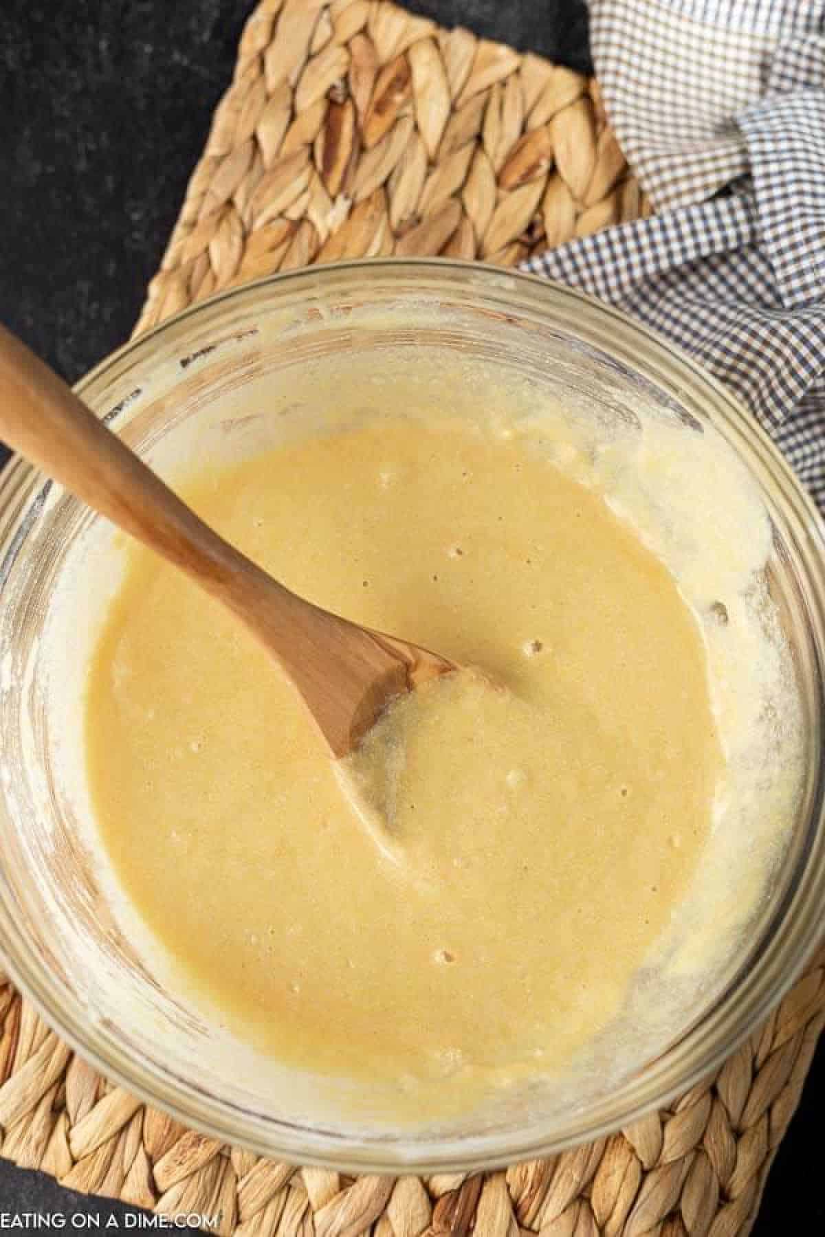 A wooden spoon rests in a glass bowl filled with smooth, light yellow batter—perfect for a honey cornbread recipe. The bowl is placed on a woven mat, and a black and white checkered cloth is partially visible next to it.