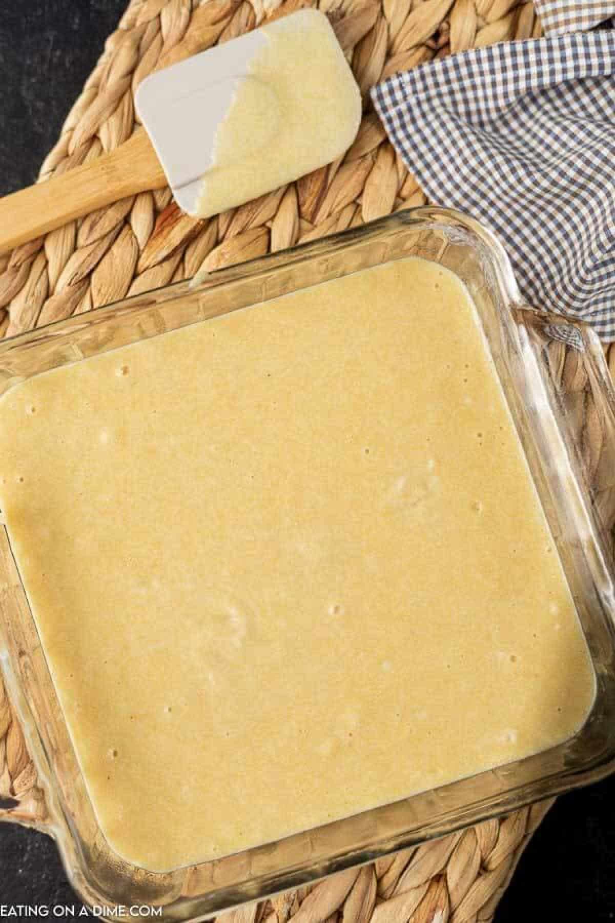 A square glass baking dish brims with uncooked honey cornbread batter, resting on a woven placemat. Nearby, a spatula coated in the sweet mixture sits atop a blue and white checkered cloth, hinting at the delicious recipe to come.