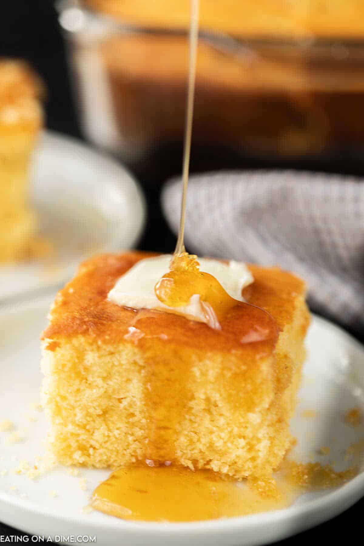 A slice of honey cornbread topped with a pat of melting butter, drizzled with golden sweetness. This delightful dessert is served on a white plate, while in the blurred background more cornbread and a checkered napkin await. Perfect for those seeking the finest honey cornbread recipe experience.