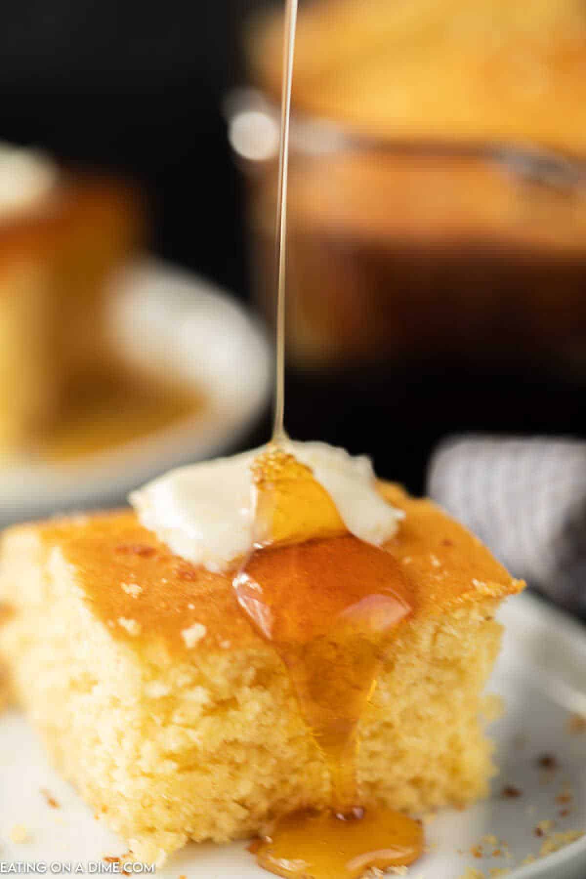 A close-up of a slice from the honey cornbread recipe with a dollop of butter, as golden honey is delicately drizzled on top. The cornbread rests on a white plate, surrounded by a blurred backdrop featuring more pieces of this delicious delight.