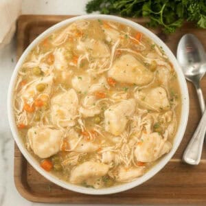 A bowl of instant pot chicken and dumplings with peas and carrots, garnished with herbs, sits on a wooden board next to a spoon and fork. Some fresh parsley is visible in the background.