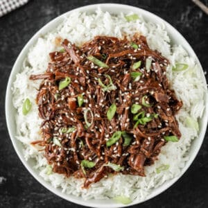 A bowl of white rice topped with shredded beef in a rich, crockpot Korean BBQ sauce, garnished with sesame seeds and chopped green onions. The dish is beautifully presented in a round bowl on a dark surface.