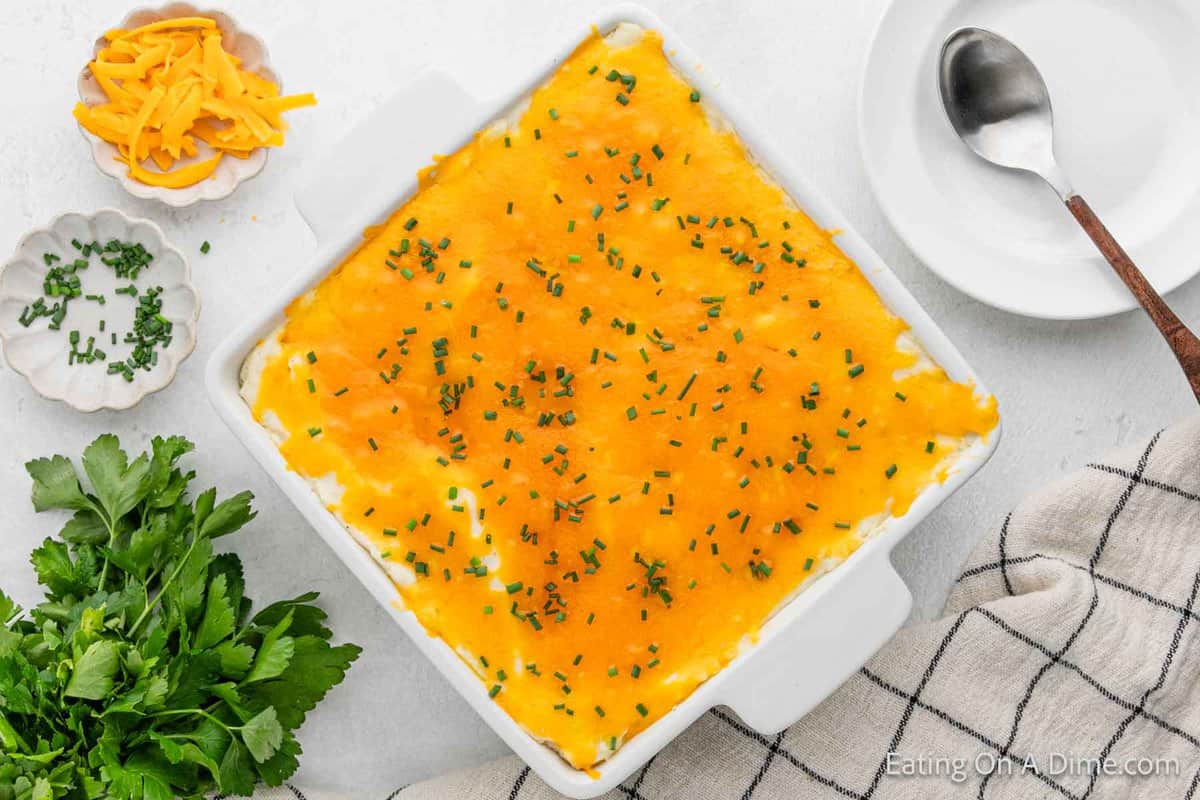 Cheesy mashed potatoes in a casserole dish with a small bowl of shredded cheese on the side with chopped chives. 