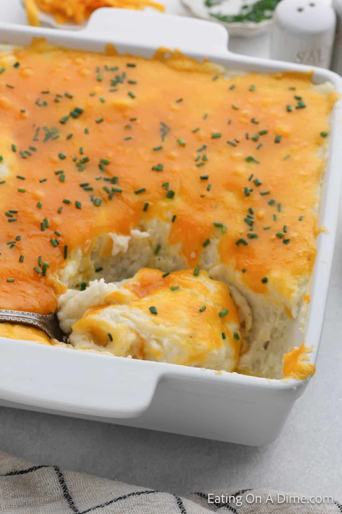 Mashed Potatoes in a casserole dish topped with melted cheddar cheese and chives with a serving on a spoon