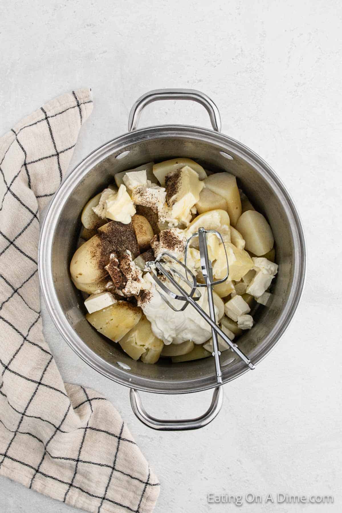 Potatoes cut into quarters in a large pot topped with seasoning, cream cheese and sour cream with hand mixer blenders