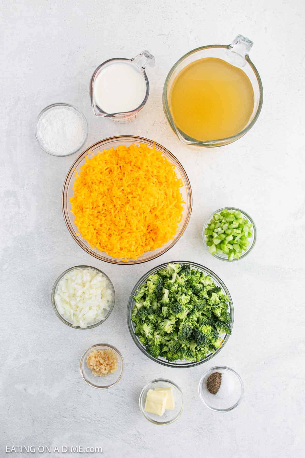 Top view of ingredients for an Instant Pot Broccoli Cheddar Soup recipe, featuring shredded cheese, diced onion, chopped broccoli, celery, minced garlic, butter, seasoning, milk, chicken broth, and flour—neatly arranged in bowls and cups on a light surface.