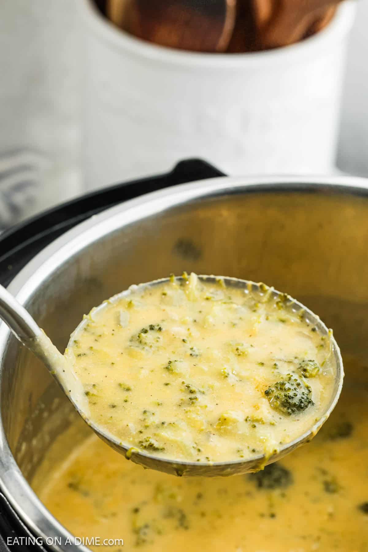 A ladle filled with Instant Pot Broccoli Cheddar Soup hovers over the pot. The soup is thick, showcasing chunks of broccoli within a cheesy texture. In the background, kitchen utensils are partially visible in a container.