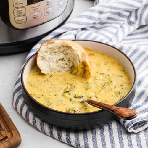 A bowl of creamy Instant Pot Broccoli Cheddar Soup topped with a slice of bread. A spoon rests inside the bowl, set on a blue and white striped cloth, while the trusty instant pot sits proudly in the background.