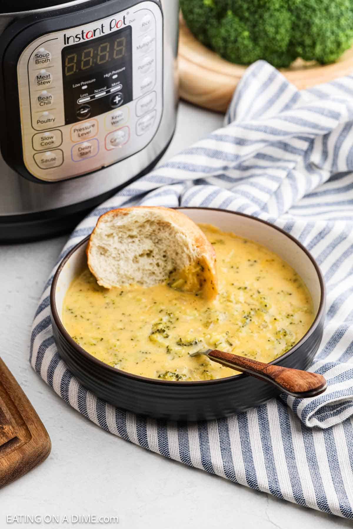 A bowl of Instant Pot Broccoli Cheddar Soup with a slice of bread sits on a striped cloth. A wooden spoon rests on the bowl's edge, while an Instant Pot stands proudly in the background next to fresh broccoli.