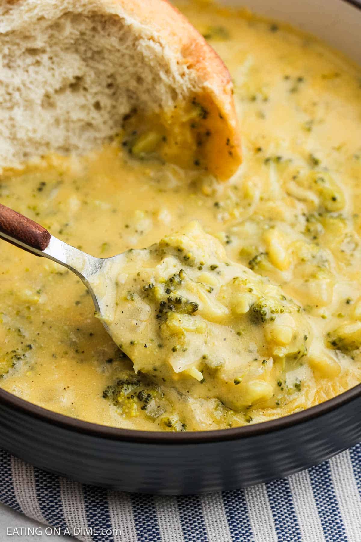 A bowl of creamy Instant Pot Broccoli Cheddar Soup topped with a slice of bread. A spoon rests in the bowl, revealing a thick portion with chunks of broccoli and melted cheese. The bowl sits on a striped fabric surface.