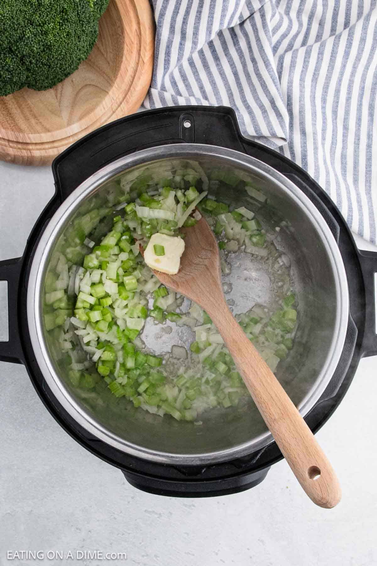 A wooden spoon stirs chopped onions and green peppers with butter in a pot, prepping the base for an Instant Pot Broccoli Cheddar Soup. The pot rests on a light-colored countertop beside a striped cloth and a wooden board adorned with fresh broccoli.