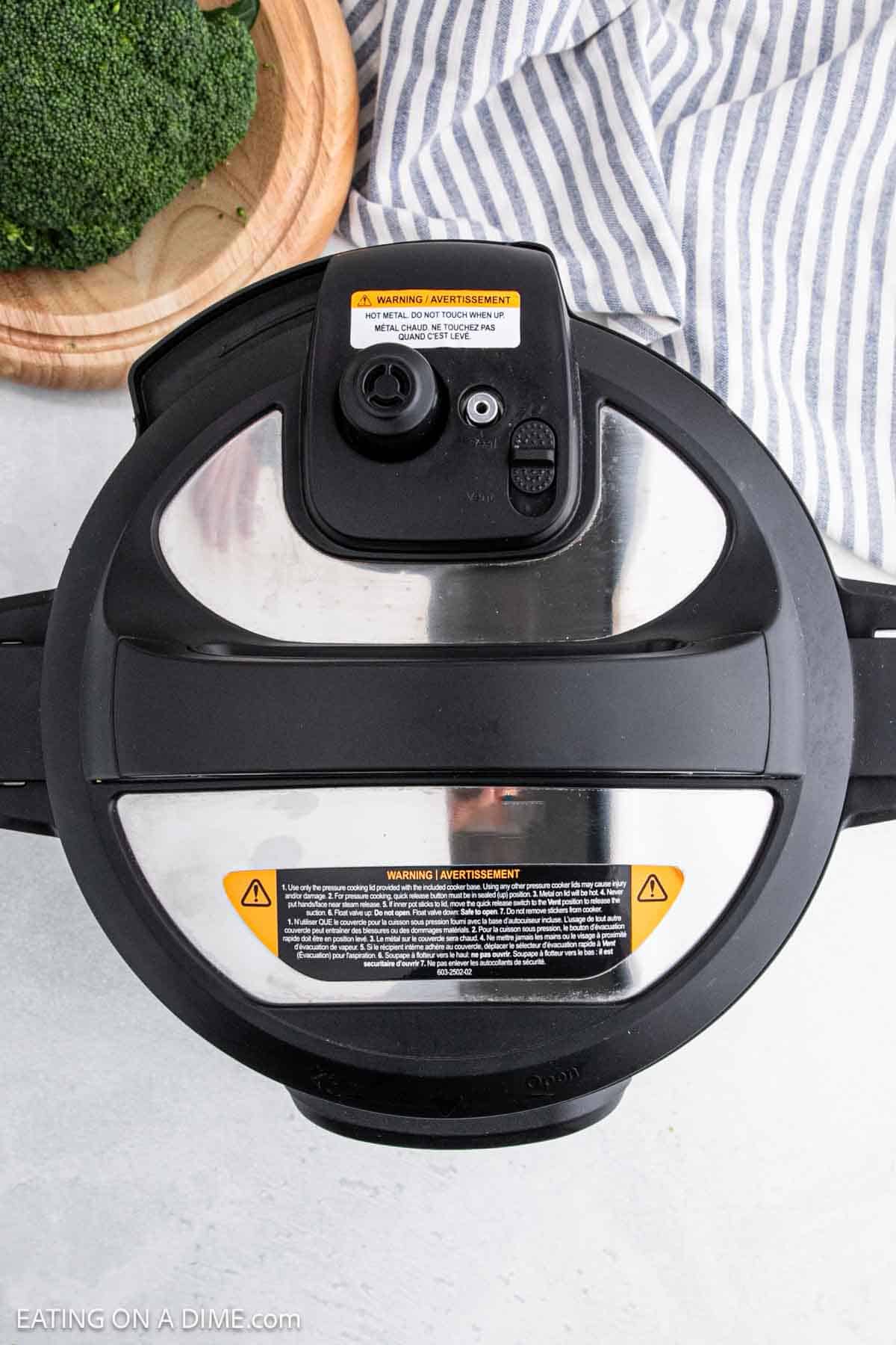 Top view of a black and stainless steel pressure cooker lid on a striped cloth, poised to create the perfect Instant Pot broccoli cheddar soup next to a wooden bowl filled with fresh broccoli.