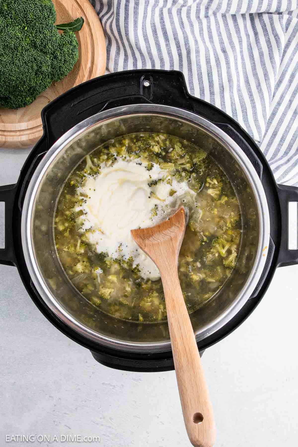 A pot of Instant Pot Broccoli Cheddar Soup is being stirred with a wooden spoon. The soup showcases broccoli pieces and a dollop of cream or yogurt on top. In the background, a wooden bowl of fresh broccoli and a striped towel add to the cozy kitchen scene.