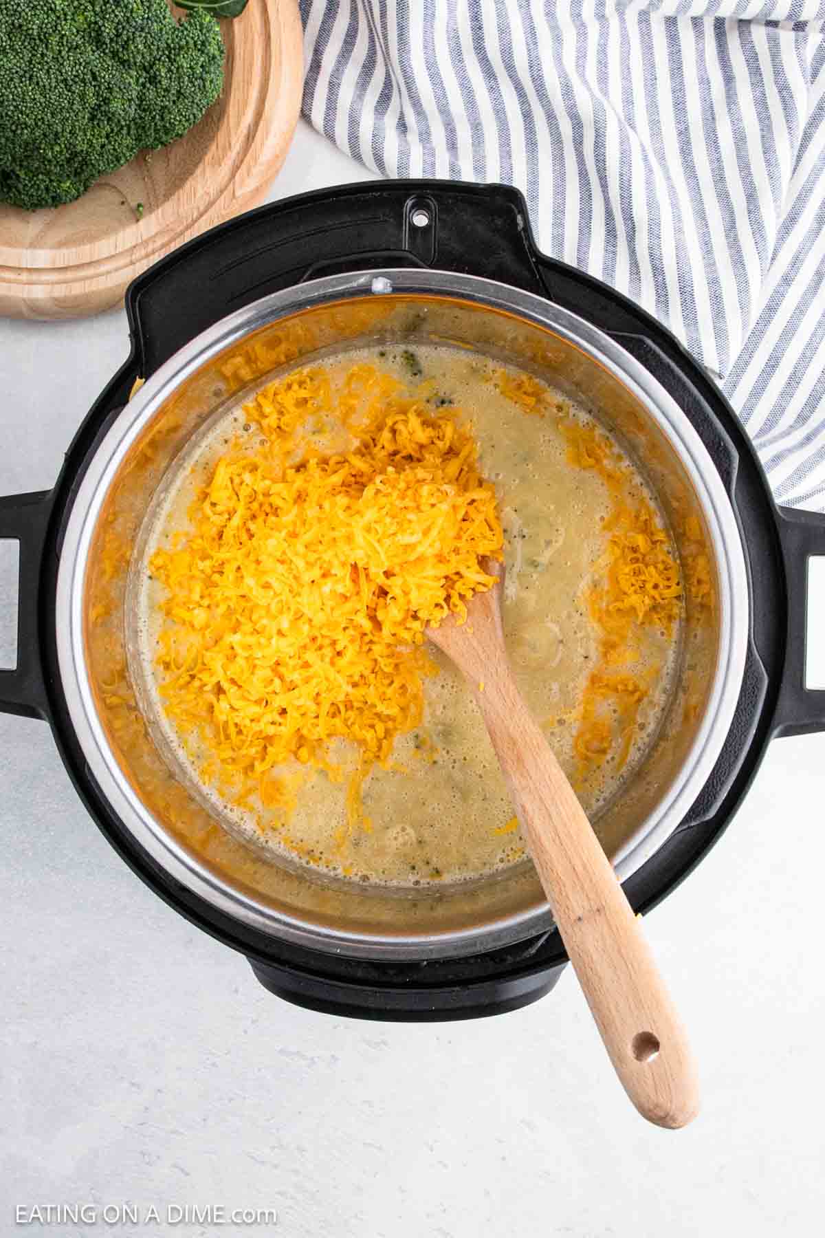 An overhead view of a pot, reminiscent of an Instant Pot, containing a mixture of melted cheese and a liquid base, being stirred with a wooden spoon. Fresh broccoli is on a nearby wooden plate, suggesting the makings of Broccoli Cheddar Soup, with a striped cloth beside it.