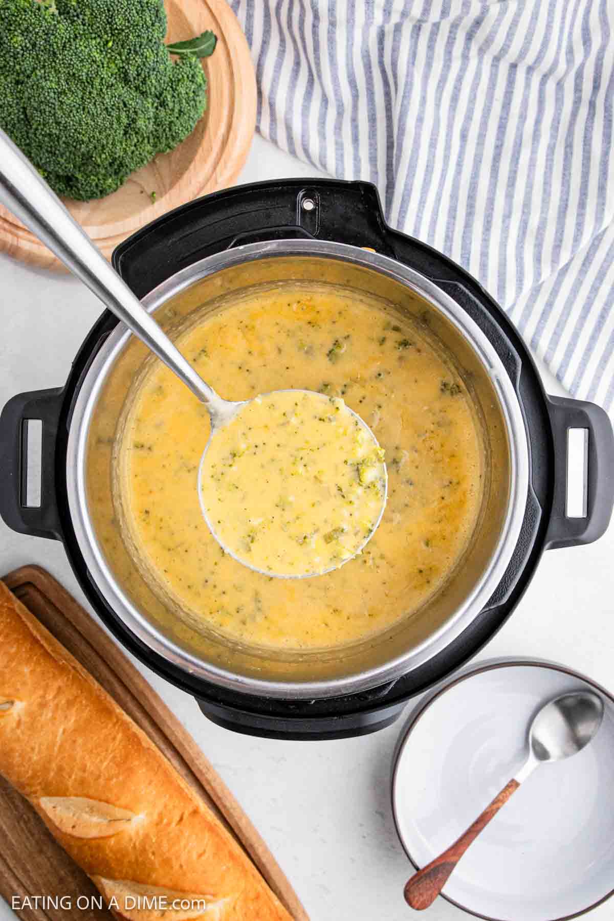 A ladle lifts the creamy goodness of Instant Pot Broccoli Cheddar Soup, surrounded by a striped cloth, a rustic loaf on a wooden board, and a white plate with a spoon. A fresh head of broccoli peeks in from the corner, completing this cozy kitchen scene.