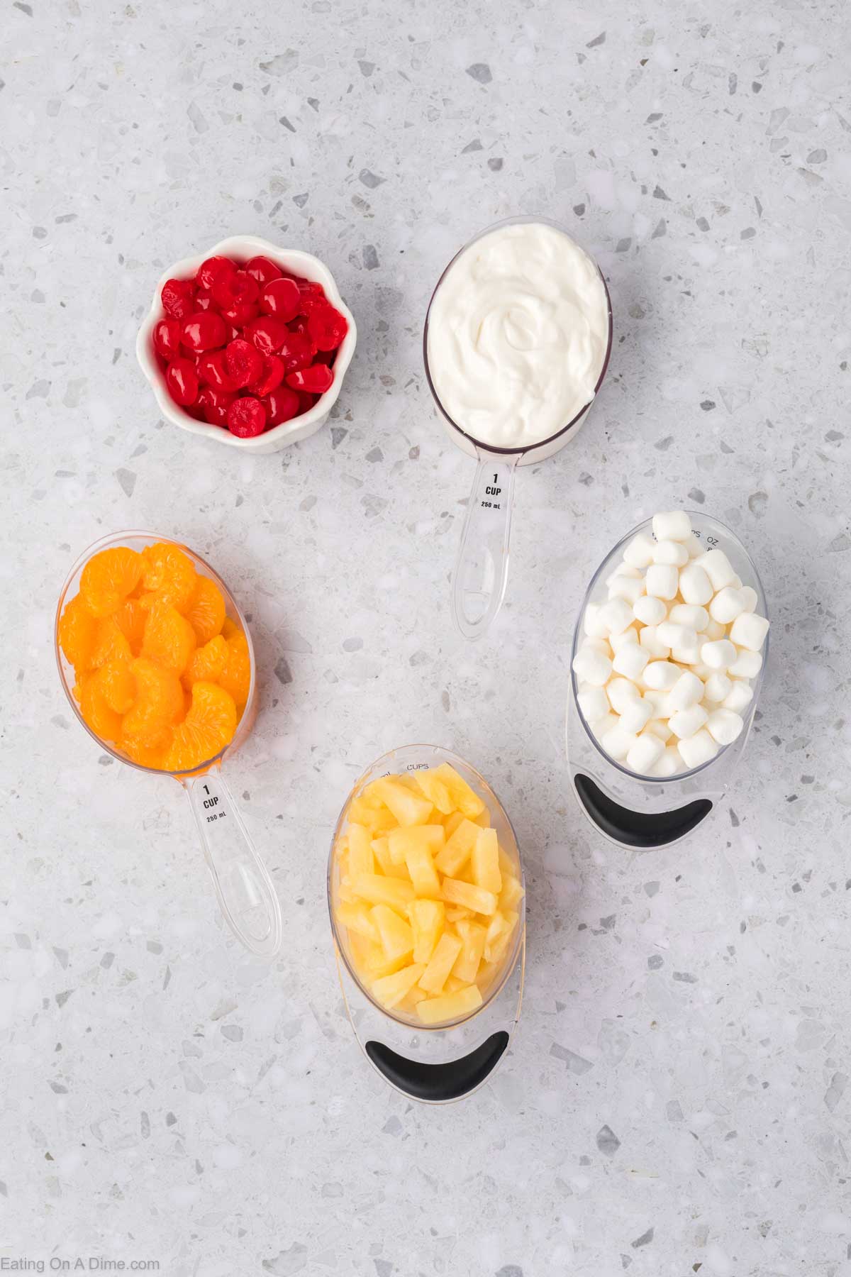 Top-down view of ingredients for a 5 Cup Salad: a bowl of red cherries, a cup of whipped cream, and measuring cups with orange segments, diced pineapples, and mini marshmallows on a speckled countertop.