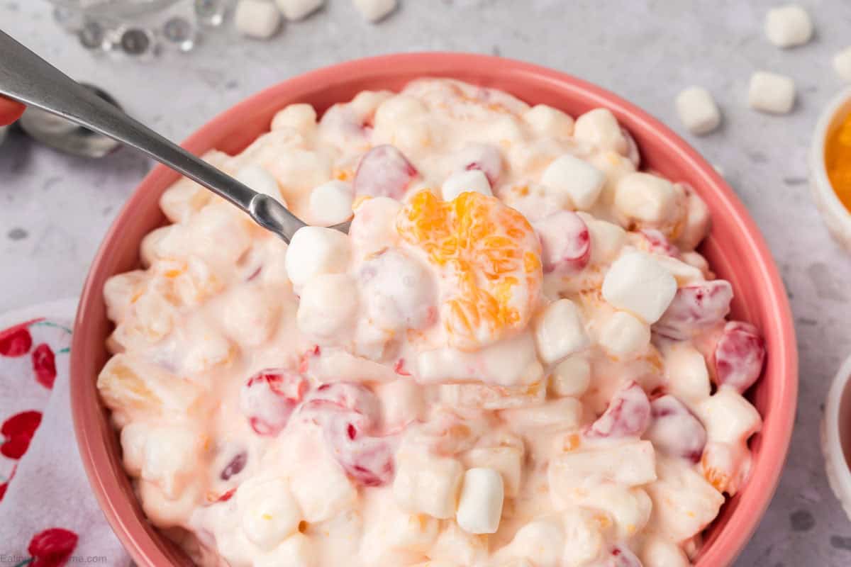 A bowl of creamy 5 Cup Salad with marshmallows, cherries, and mandarin orange slices. A spoon lifts a portion of the salad, showcasing its colorful and textured ingredients. The bowl rests on a light gray surface.