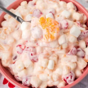 A close-up of a bowl filled with 5 Cup Salad showcases a creamy fruit medley, featuring mini marshmallows, orange segments, and red cherries. A spoon lifts a portion, highlighting the delightful mix of marshmallows and fruit in the smooth mixture.