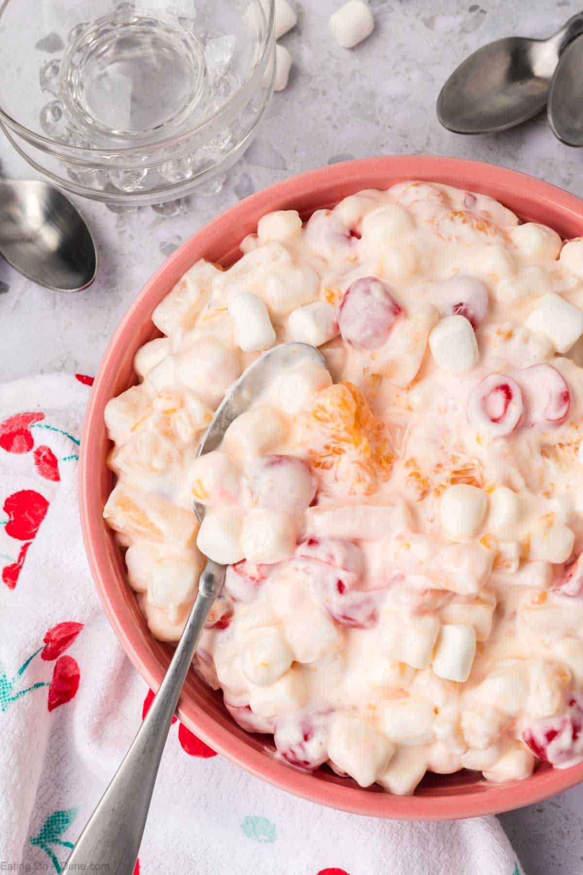 A pink bowl brimming with 5 Cup Salad features a fruity mix of marshmallows, mandarin oranges, and red cherries in a creamy sauce. A spoon rests inside the bowl, which is set on a floral-patterned cloth surrounded by extra spoons and a glass.