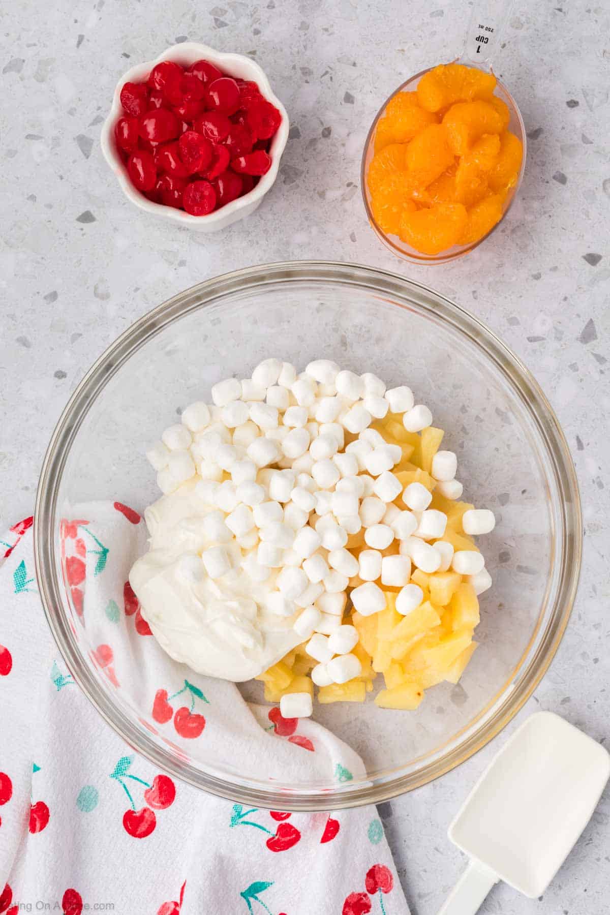 A glass bowl brimming with 5 Cup Salad, featuring marshmallows, cream, and pineapple chunks. Nearby are small bowls of red cherries and mandarin oranges. A cherry-patterned towel and a white spatula rest on the speckled countertop.