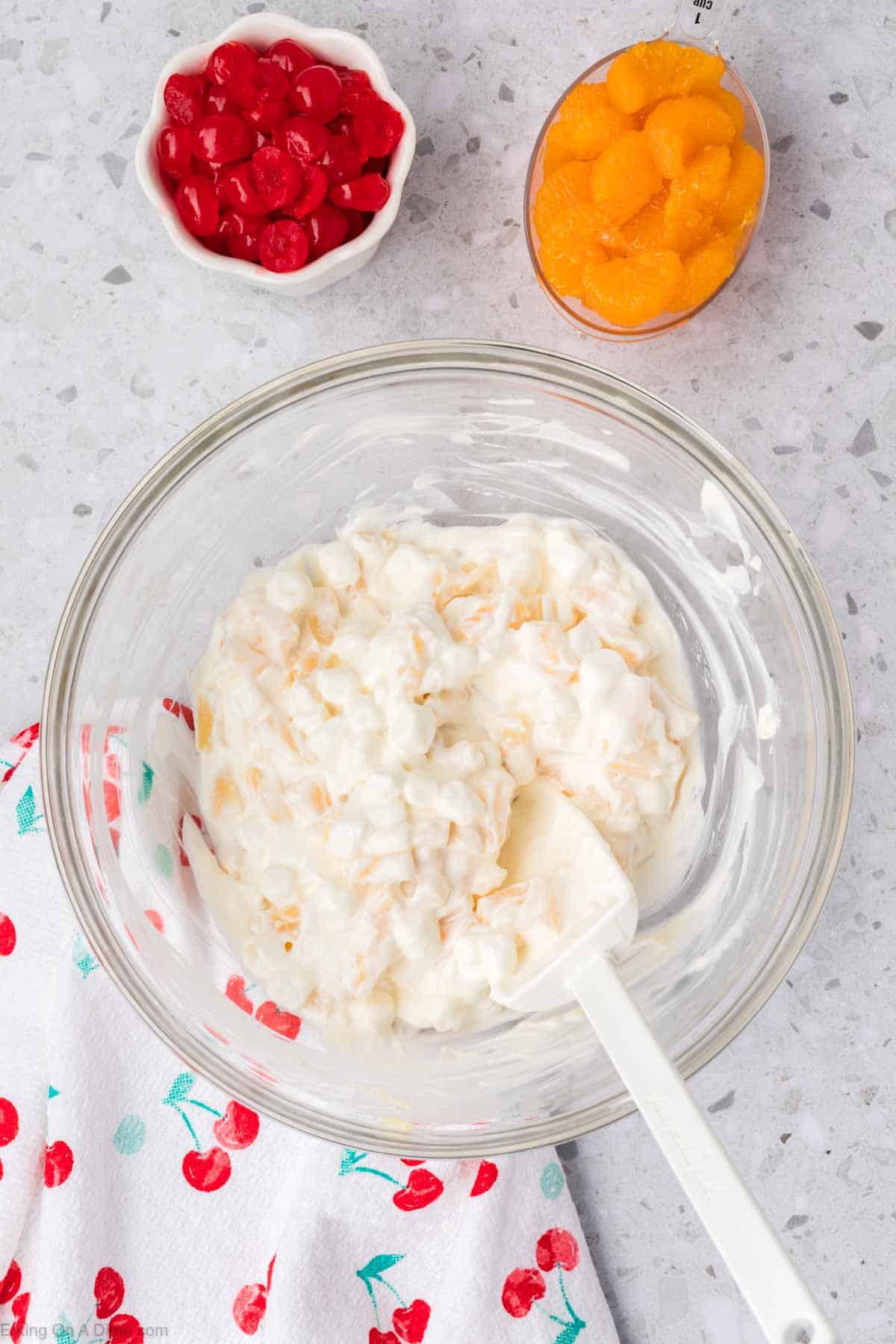 A glass bowl with creamy 5 Cup Salad mixture and a spatula inside. Nearby are small bowls of red cherries and orange mandarin slices. The setting includes a cherry-patterned white cloth on a speckled countertop, adding charm to this delightful scene.