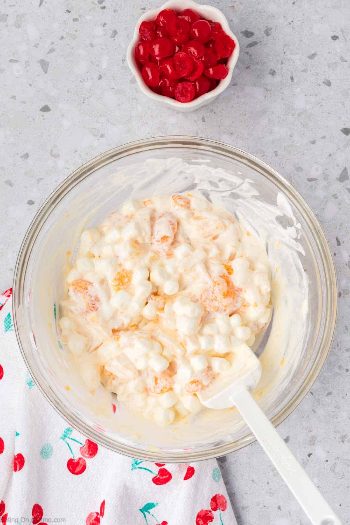 A glass bowl holds a 5 Cup Salad, a creamy dessert mixture of mini marshmallows and mandarin oranges, on a speckled countertop. Nearby, a small bowl of red maraschino cherries rests beside cherry-patterned cloth.