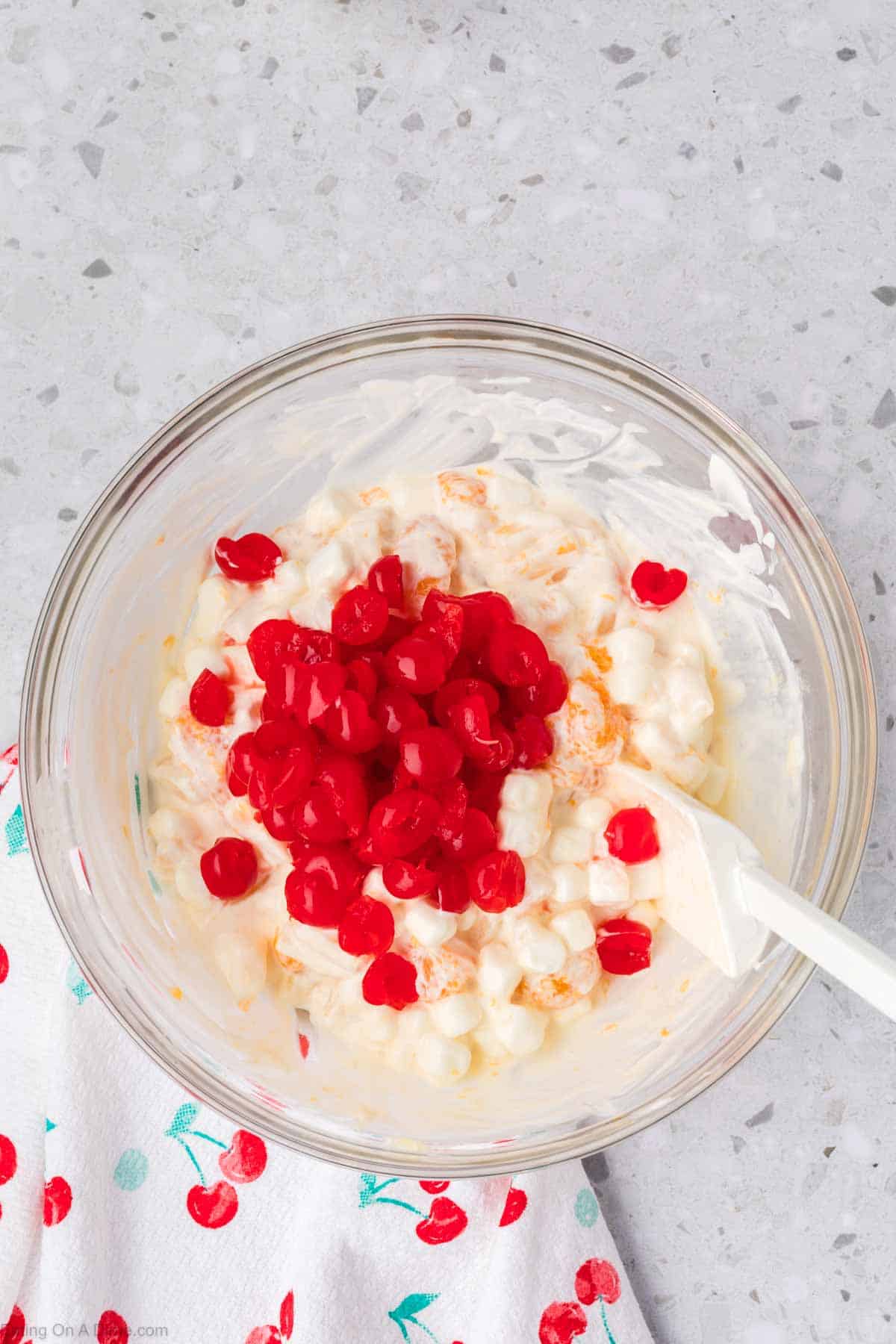 A glass bowl on a gray countertop holds a delightful 5 Cup Salad, featuring whipped cream, juicy oranges, and small marshmallows, all crowned with red cherries. A white spatula rests in the bowl, with a cherry-patterned cloth peeking out from beneath.