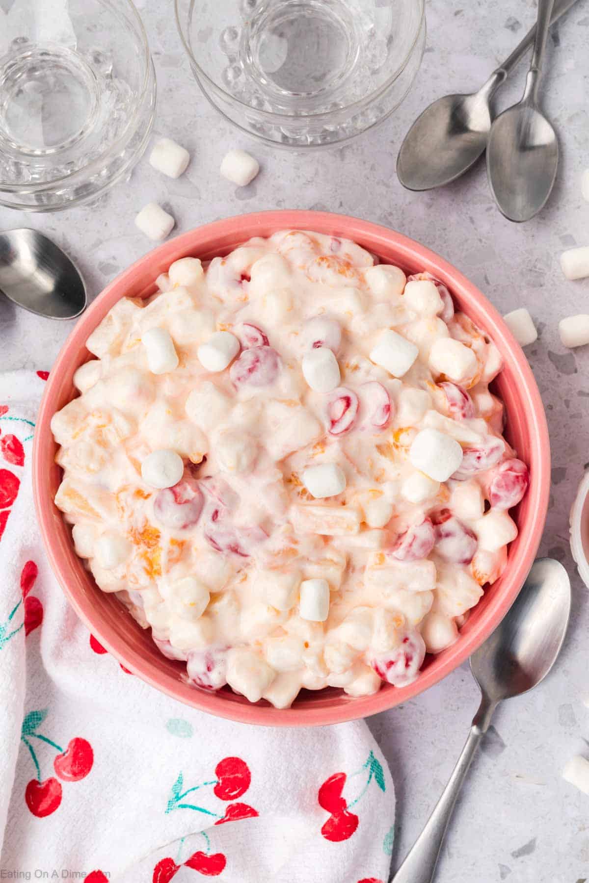 A delightful 5 Cup Salad featuring a creamy fruit blend with marshmallows, cherries, and mandarin segments. The dish is elegantly presented amid empty glass bowls, spoons, and a cherry-patterned cloth on a light surface.