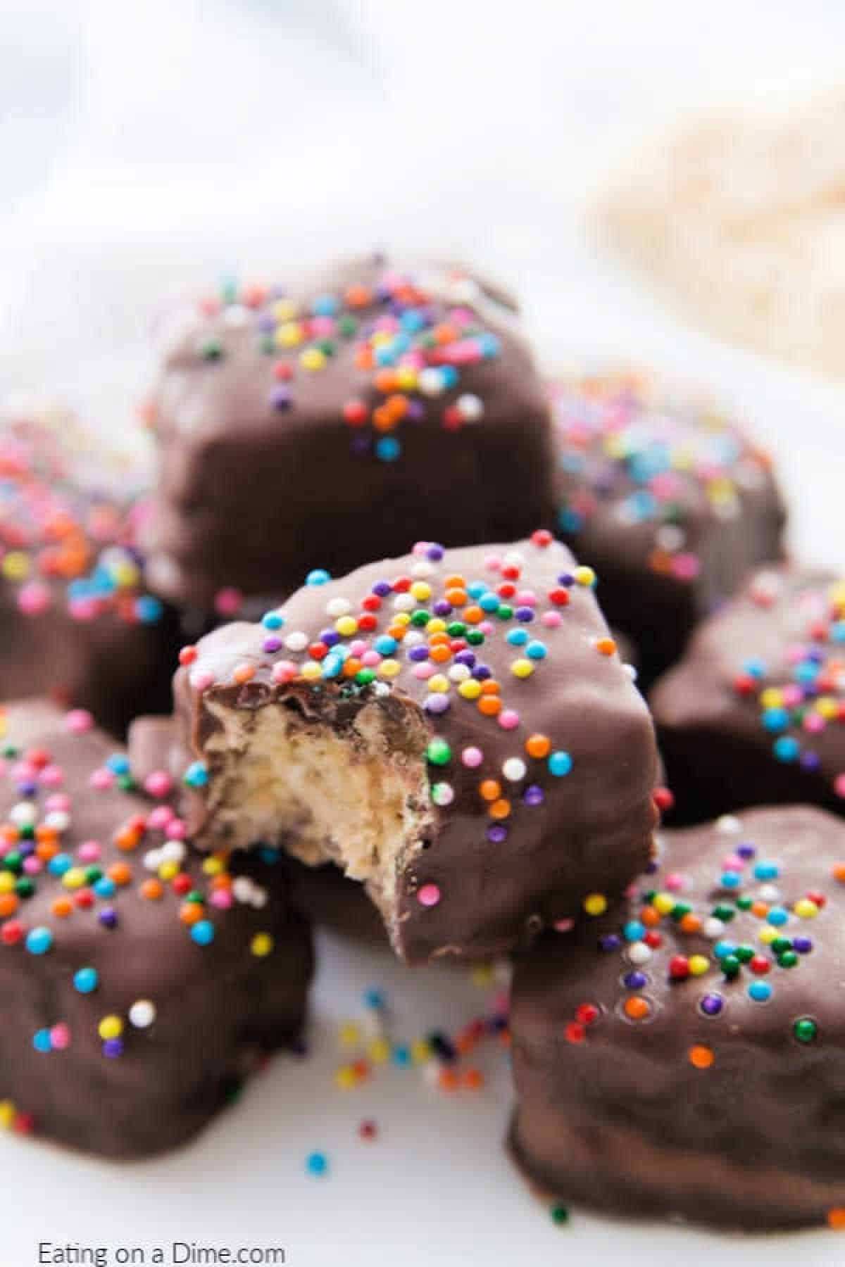 A close-up of chocolate-covered rice krispie treats topped with colorful rainbow sprinkles. One treat has a bite taken out, revealing a creamy filling. They are arranged on a white surface.