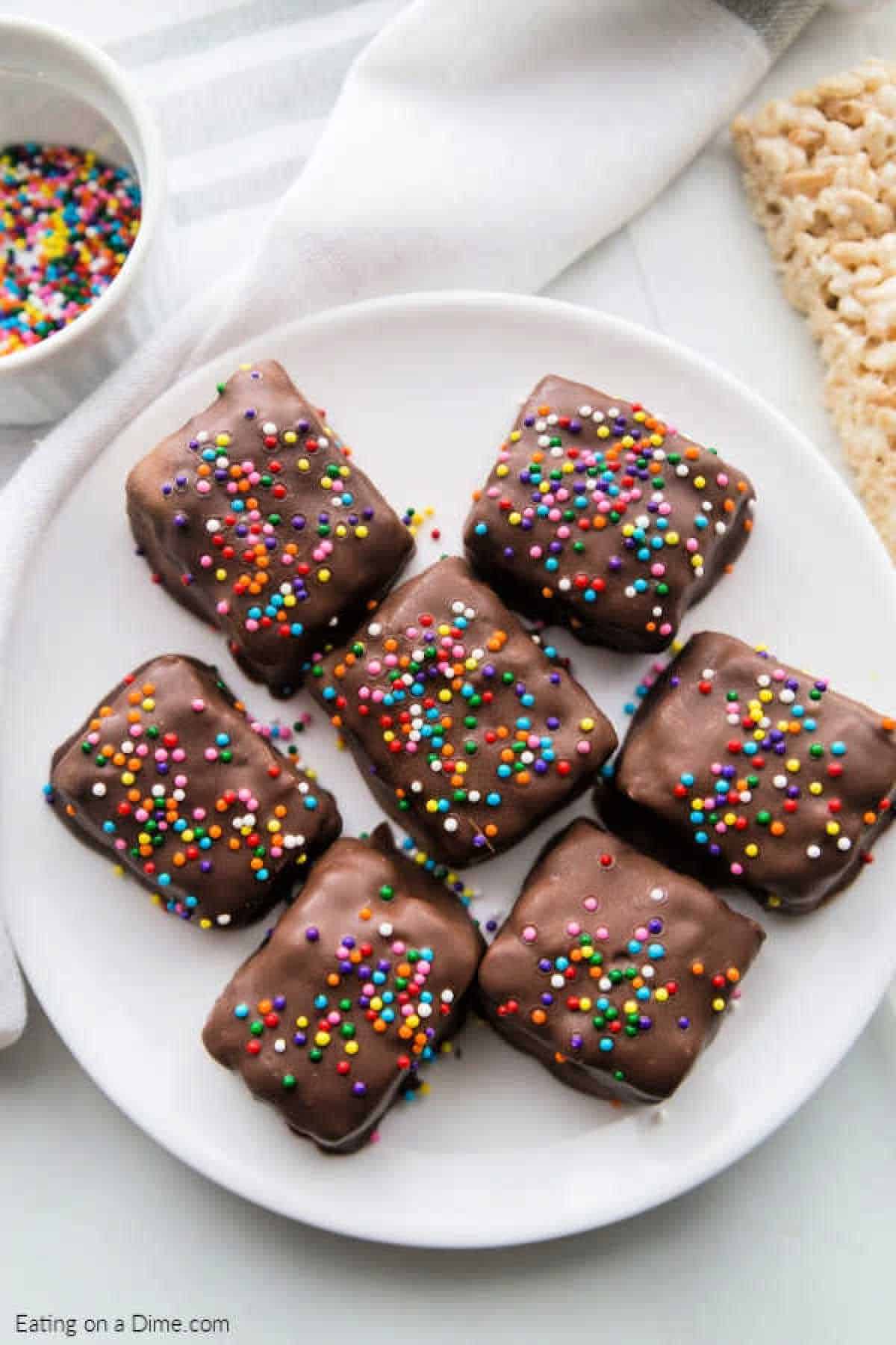 A white plate displays eight chocolate-covered Rice Krispie treats, each adorned with colorful sprinkles. In the background, a white bowl brims with additional sprinkles, nestled beside a crisp white napkin and an extra treat.