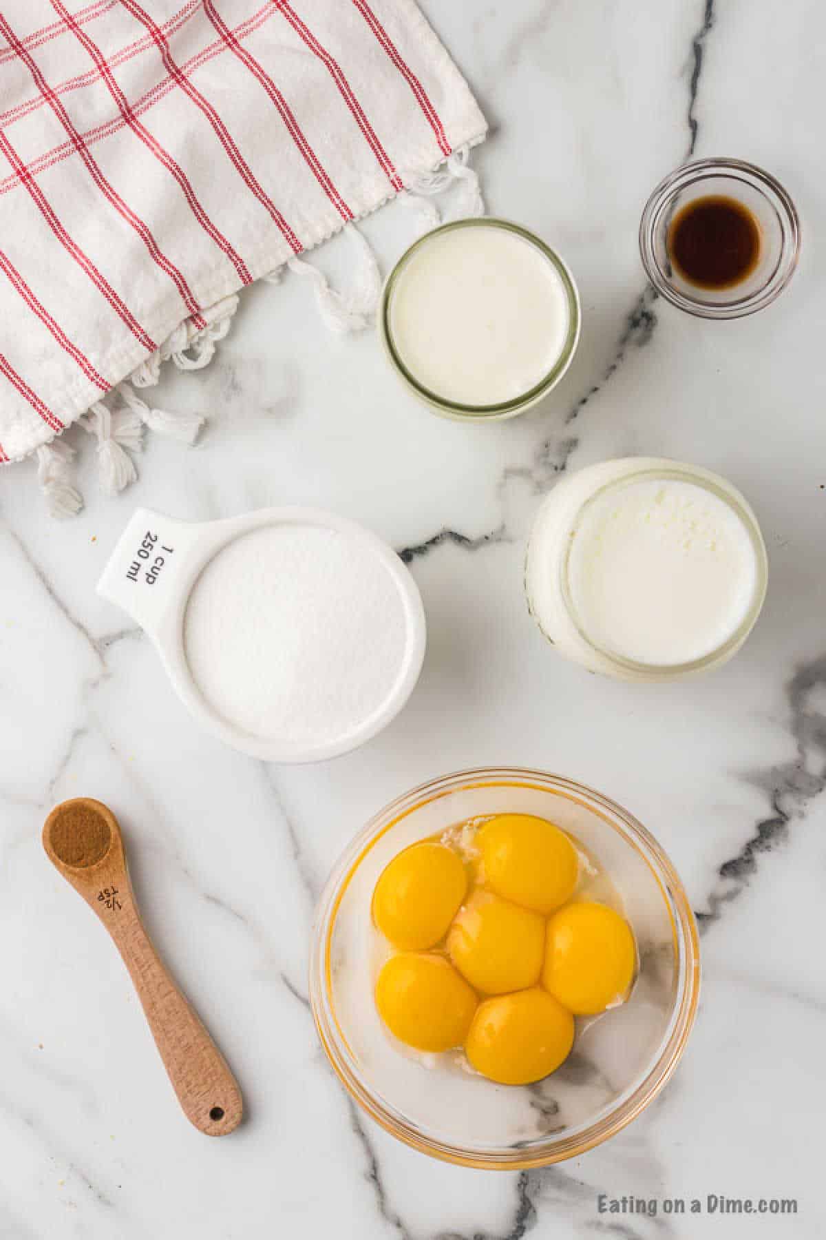 A marble countertop displays various ingredients for cooking a homemade eggnog recipe. There are six egg yolks in a glass bowl, a wooden spoon with a spice, a cup labeled "1 cup sugar" containing sugar, two small glass containers with liquid, a small glass jar with a dark liquid, and a red-striped cloth.