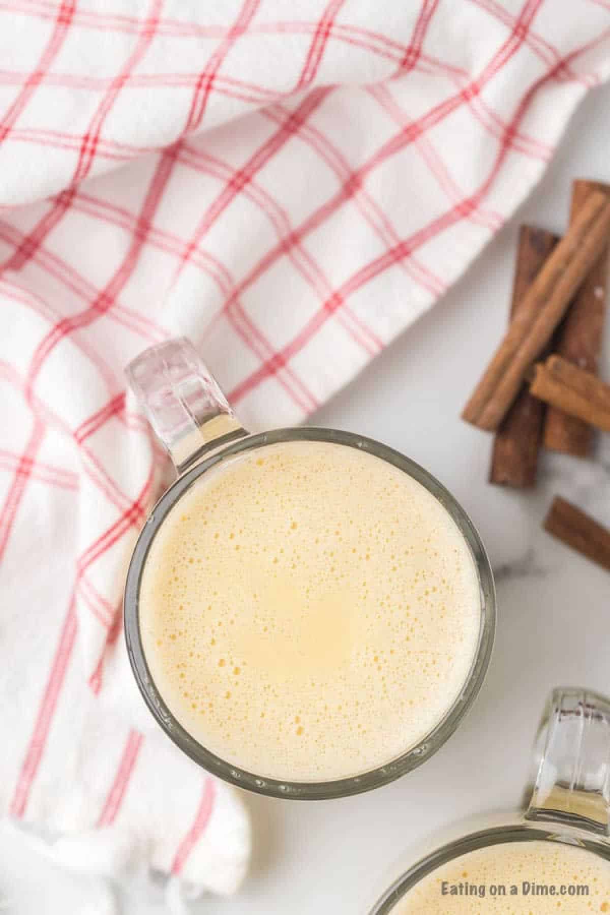 A clear mug filled with frothy, creamy eggnog made from a homemade eggnog recipe sits on a white table next to a red and white checkered cloth. Cinnamon sticks are placed nearby, adding a warm and cozy feel to the setting.