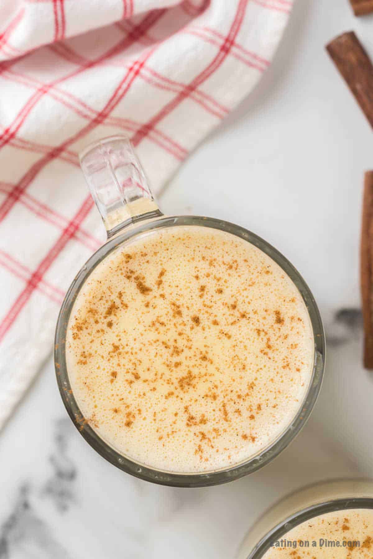 A glass mug filled with frothy eggnog, made from a homemade eggnog recipe and sprinkled with ground nutmeg, sits on a white marble countertop. Beside it is a red and white checkered cloth, and part of a cinnamon stick is visible on the right side of the image.