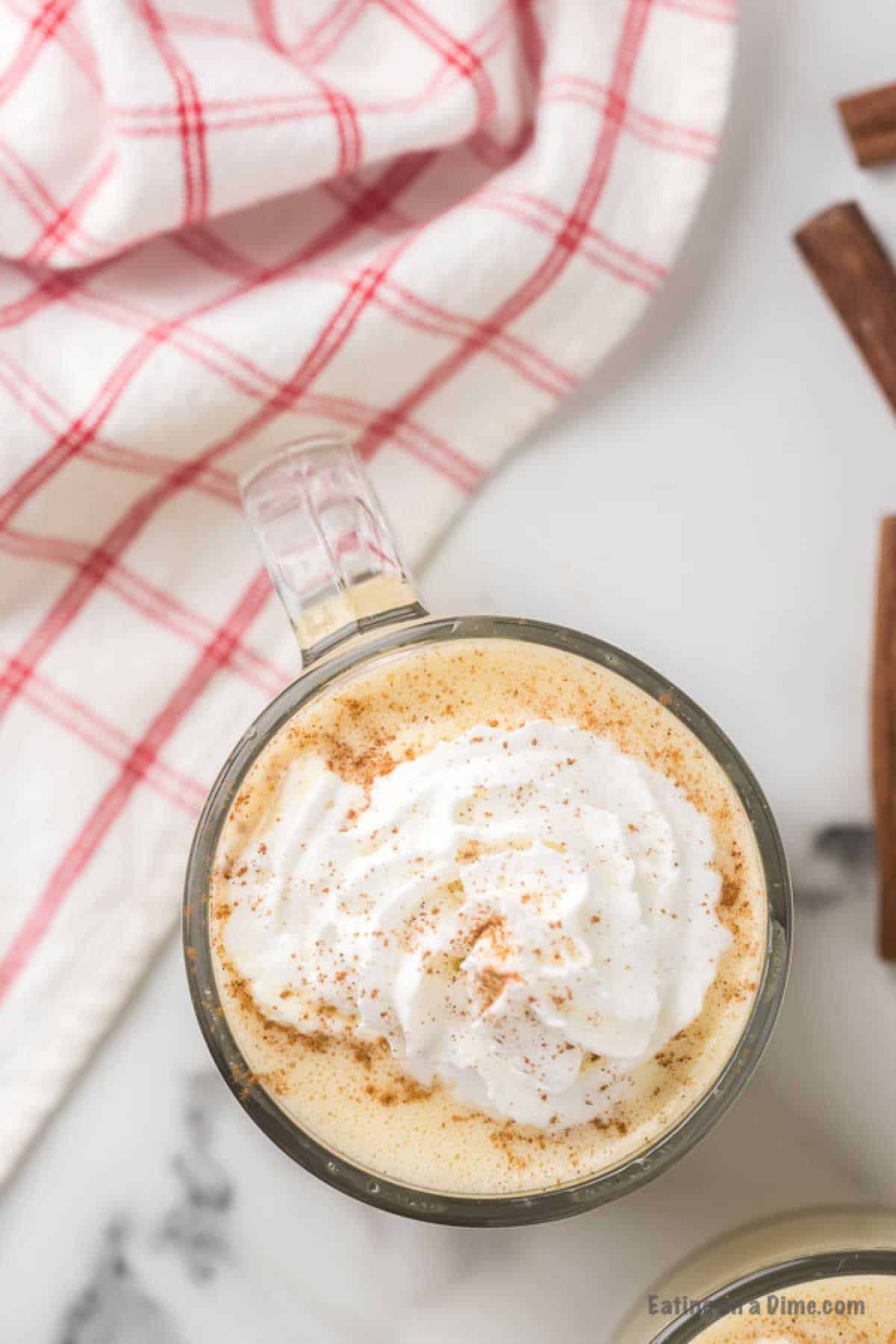 A glass mug filled with a creamy beverage topped with whipped cream and sprinkled with a bit of cinnamon, reminiscent of a homemade eggnog recipe. The mug is placed on a white marble surface next to a couple of cinnamon sticks and a red and white checkered cloth.