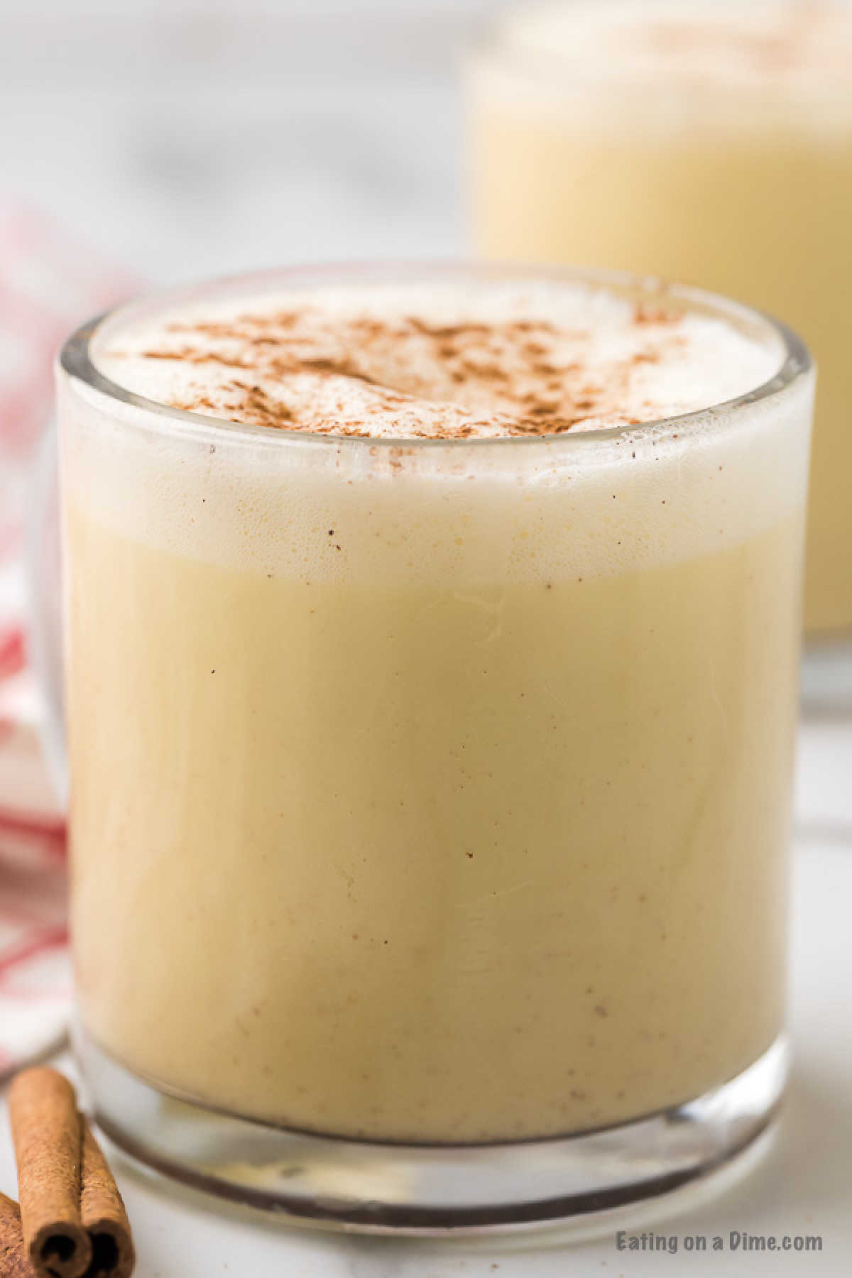 A glass filled with creamy homemade eggnog, topped with a sprinkle of ground cinnamon. There is a cinnamon stick placed beside the glass. The background is blurred, showing another glass of eggnog. The photo has a cozy and festive ambiance.