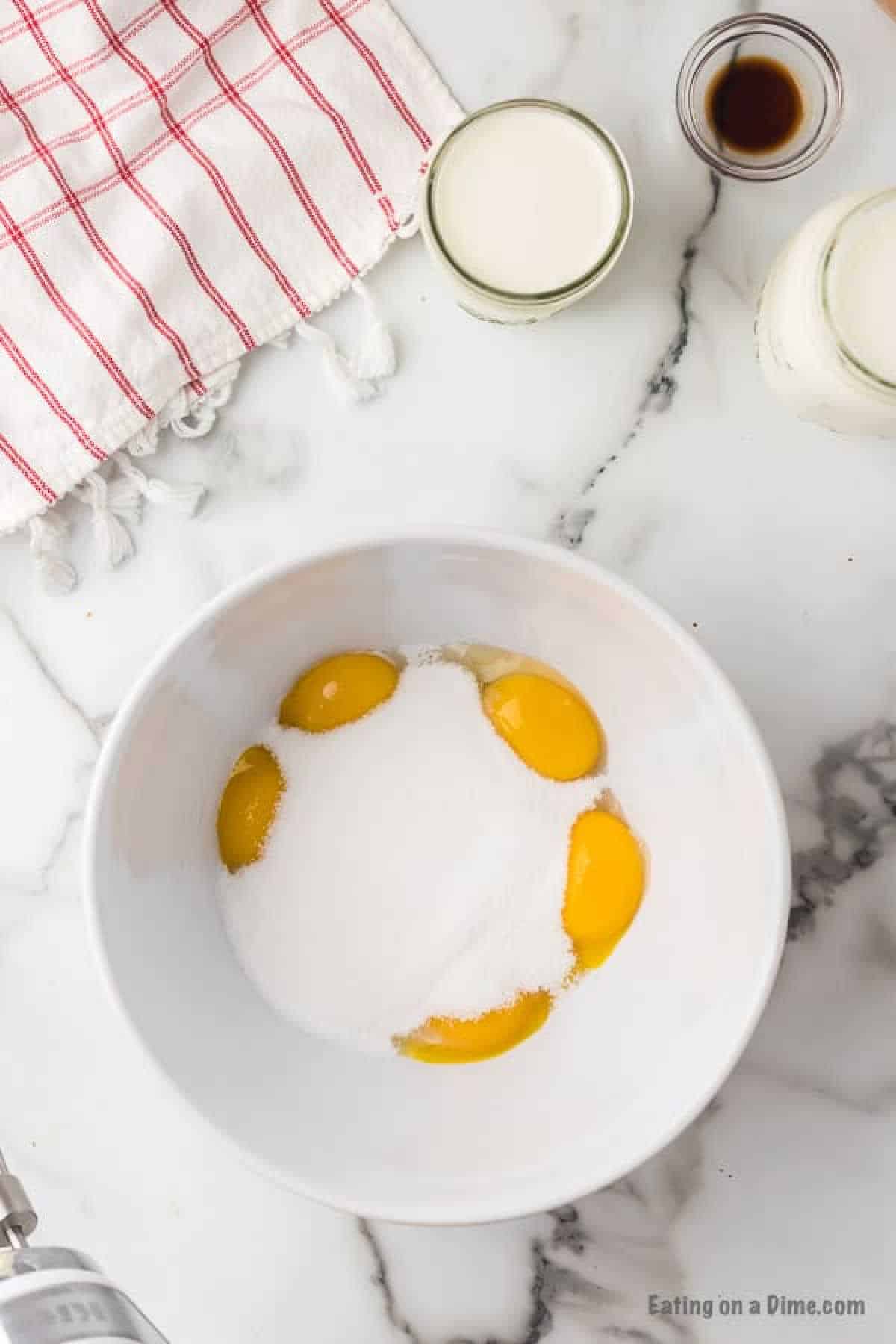 A white bowl with four egg yolks and a mound of sugar sits on a marble countertop. Surrounding the bowl are a red and white striped dish towel, a glass of milk, a small glass of vanilla extract, and a jar labeled cream—essentials for crafting your homemade eggnog recipe.