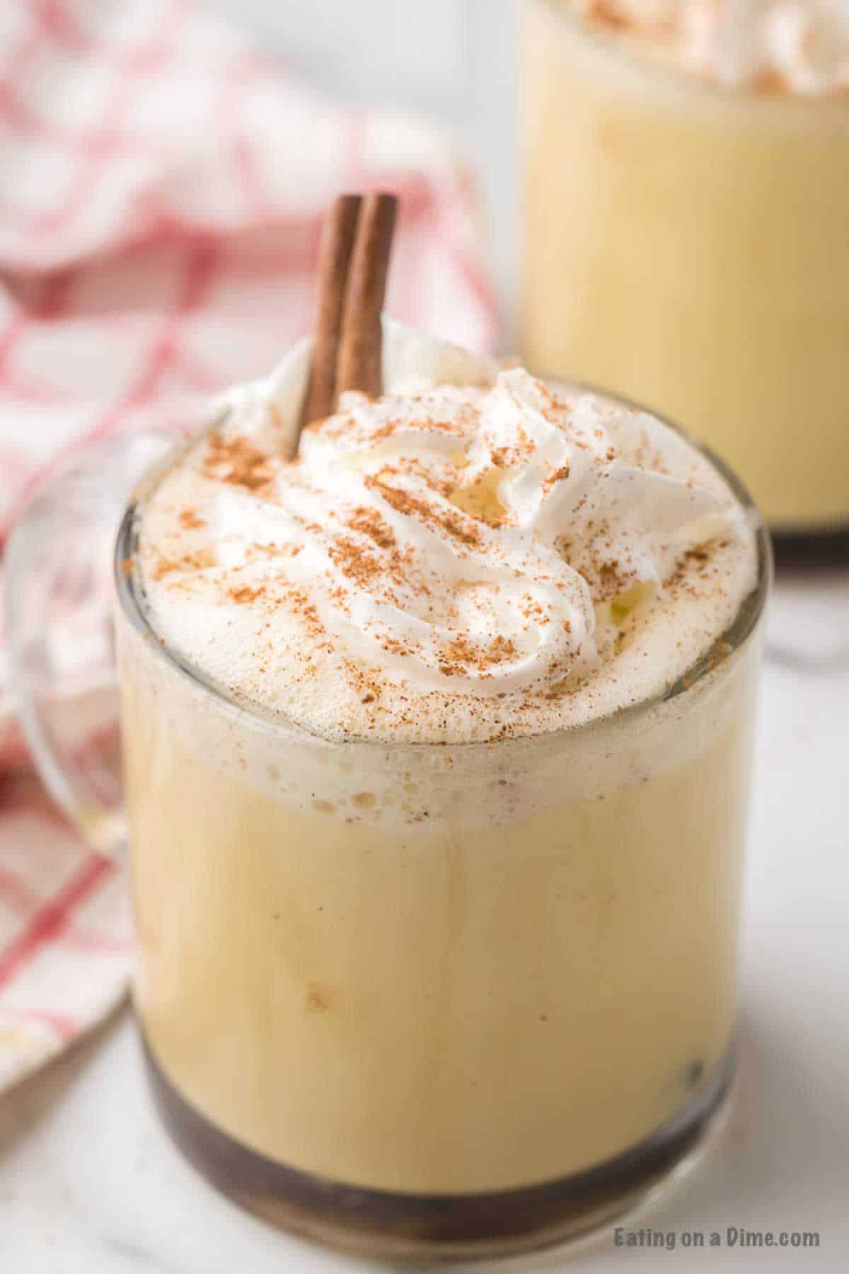 A clear glass mug filled with creamy homemade eggnog, topped with whipped cream and sprinkled with cinnamon, sits on a white surface. A cinnamon stick is inserted into the drink. A red and white checkered cloth is in the background, partially visible.