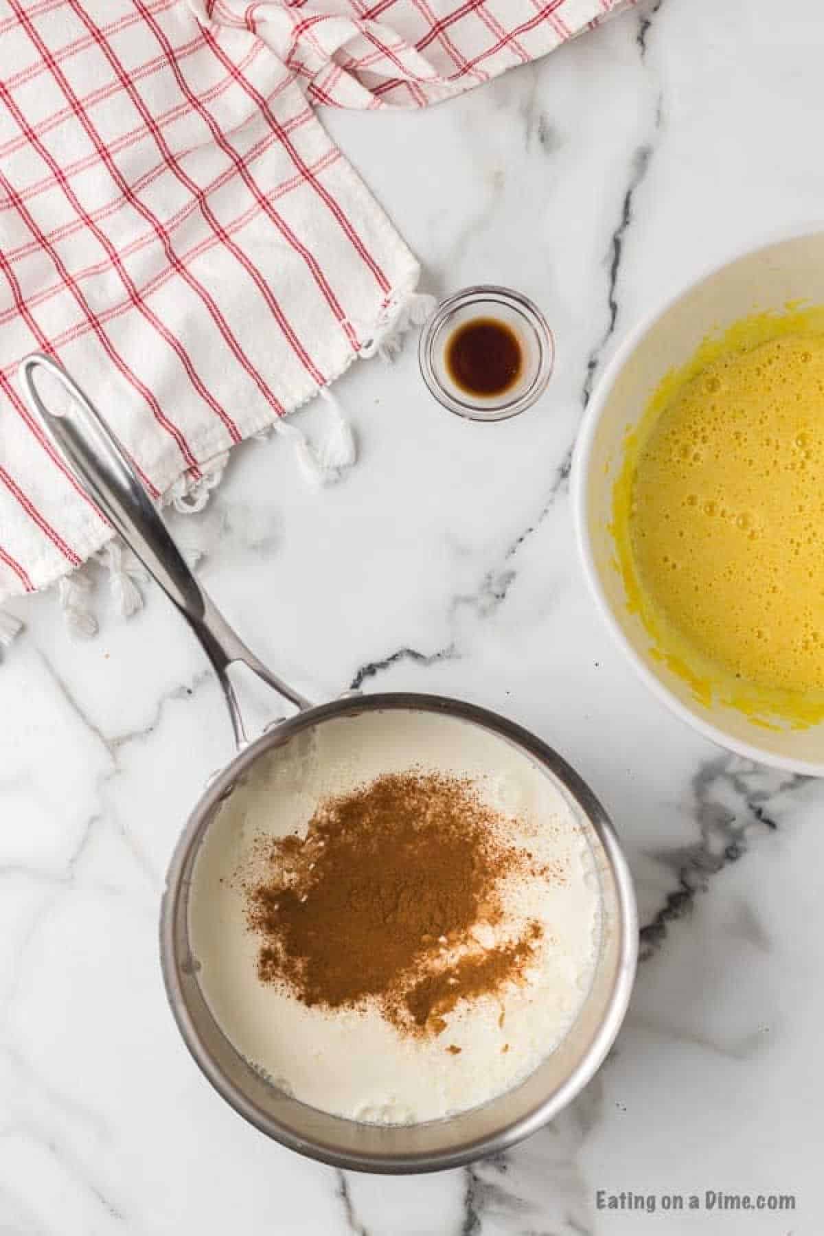 A kitchen counter with ingredients for a homemade eggnog recipe. A bowl of beaten eggs is visible on the right, a saucepan with cream and spices on the left, and a small dish of vanilla extract above. A red and white striped cloth is partially seen in the top left corner.