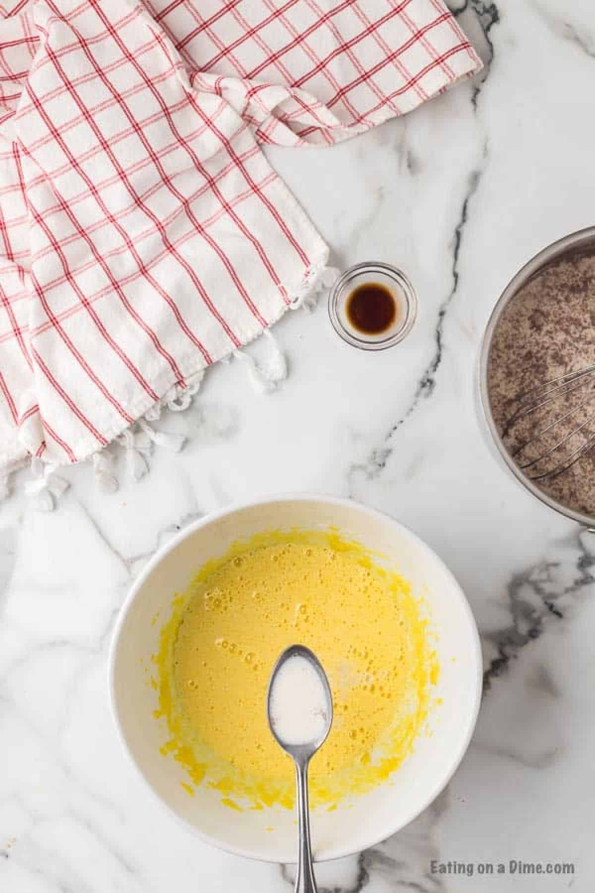 A mixing bowl with a beaten yellow mixture and a spoon, next to a smaller bowl with a brown powdery substance and a whisk, a small dish of vanilla extract, and a red and white checkered cloth on a marble surface—all the essentials for your homemade eggnog recipe.