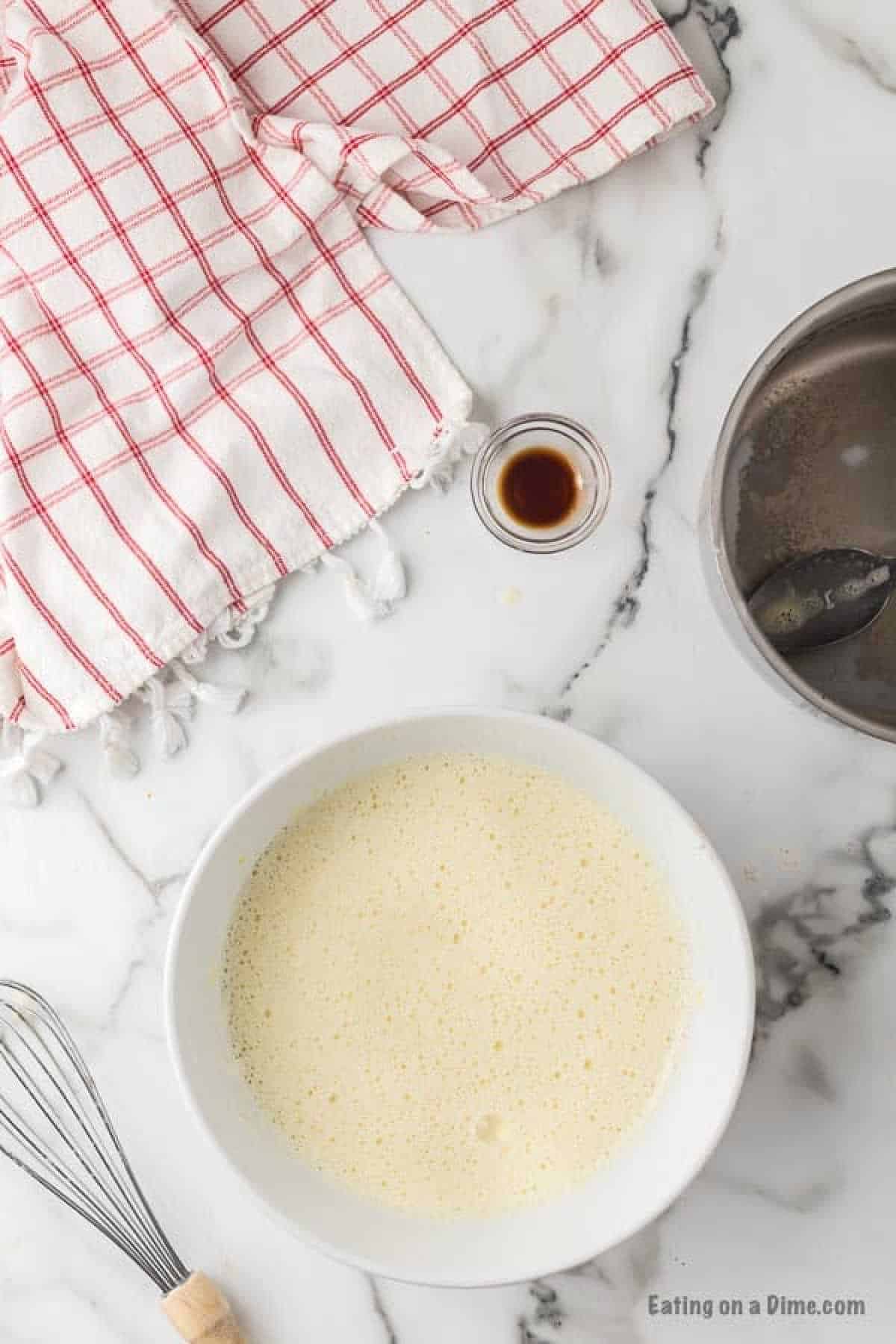 A white bowl filled with a frothy, light-colored mixture is placed on a marble countertop. Nearby are a whisk, a small glass with dark liquid, a red and white checkered cloth, and a metal pot with a spoon—perfect essentials for crafting your homemade eggnog recipe.