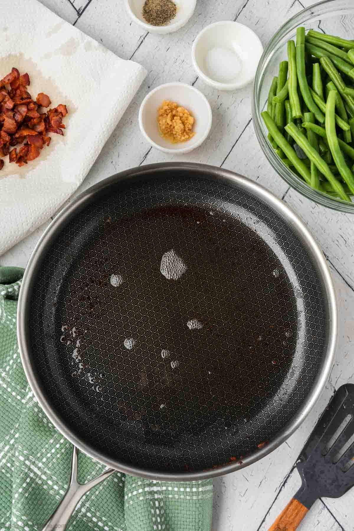 Bacon grease oil in a skillet with small bowls of minced garlic, salt, pepper, and a bowl of fresh green beans and a plate of cooked chopped bacon