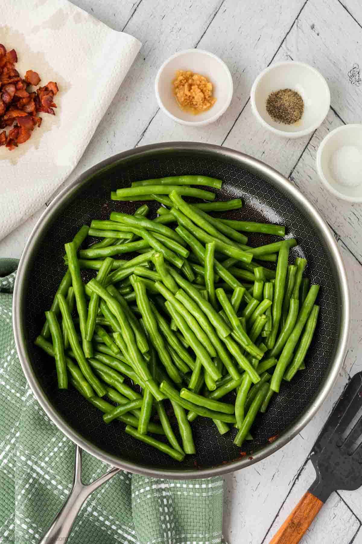Fresh green beans in a skillet with small bowls of minced garlic, pepper, salt and a plate lined with paper towel with cooked chopped bacon