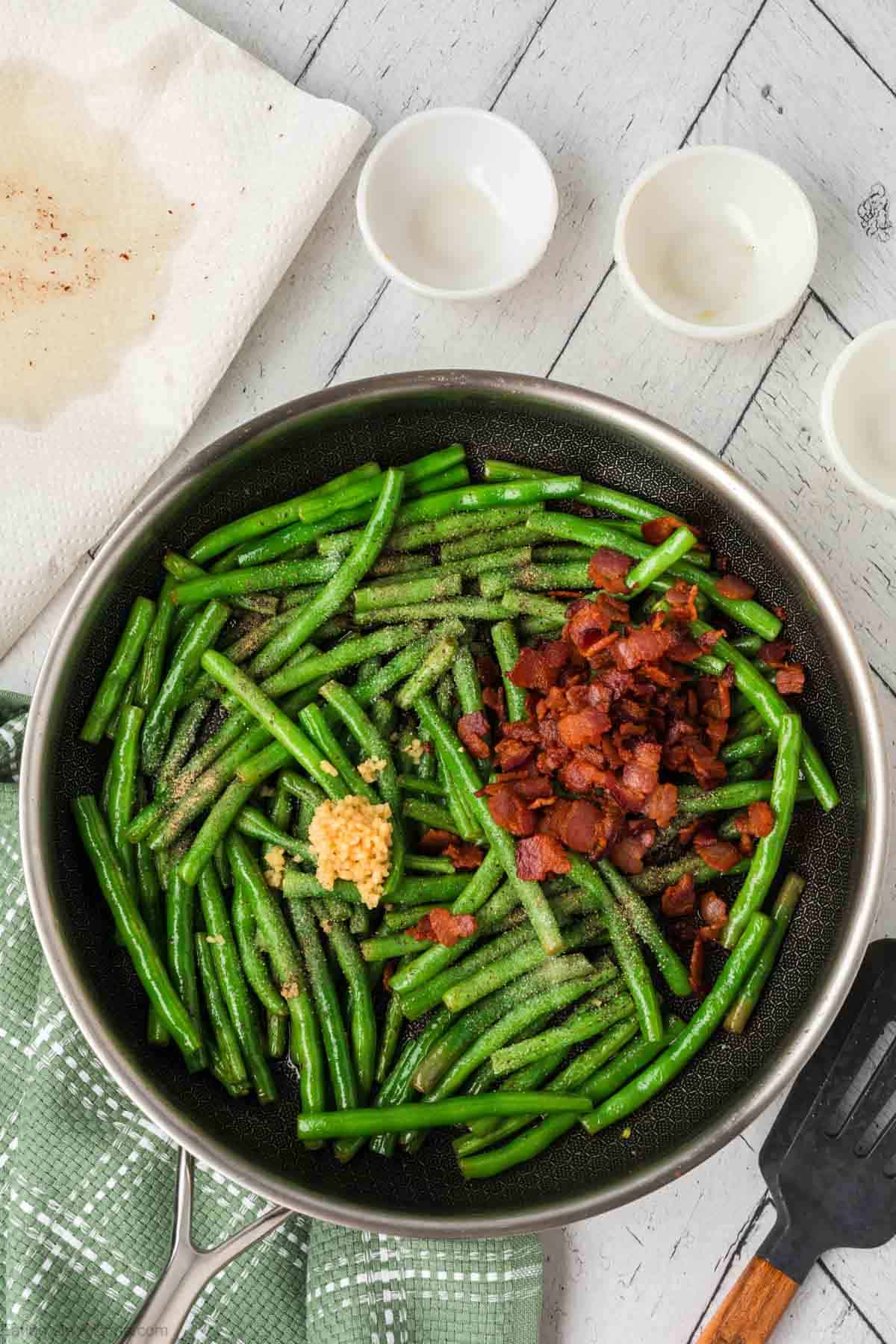 Fresh green beans in a skillet topped with minced garlic, chopped cooked bacon and seasoning