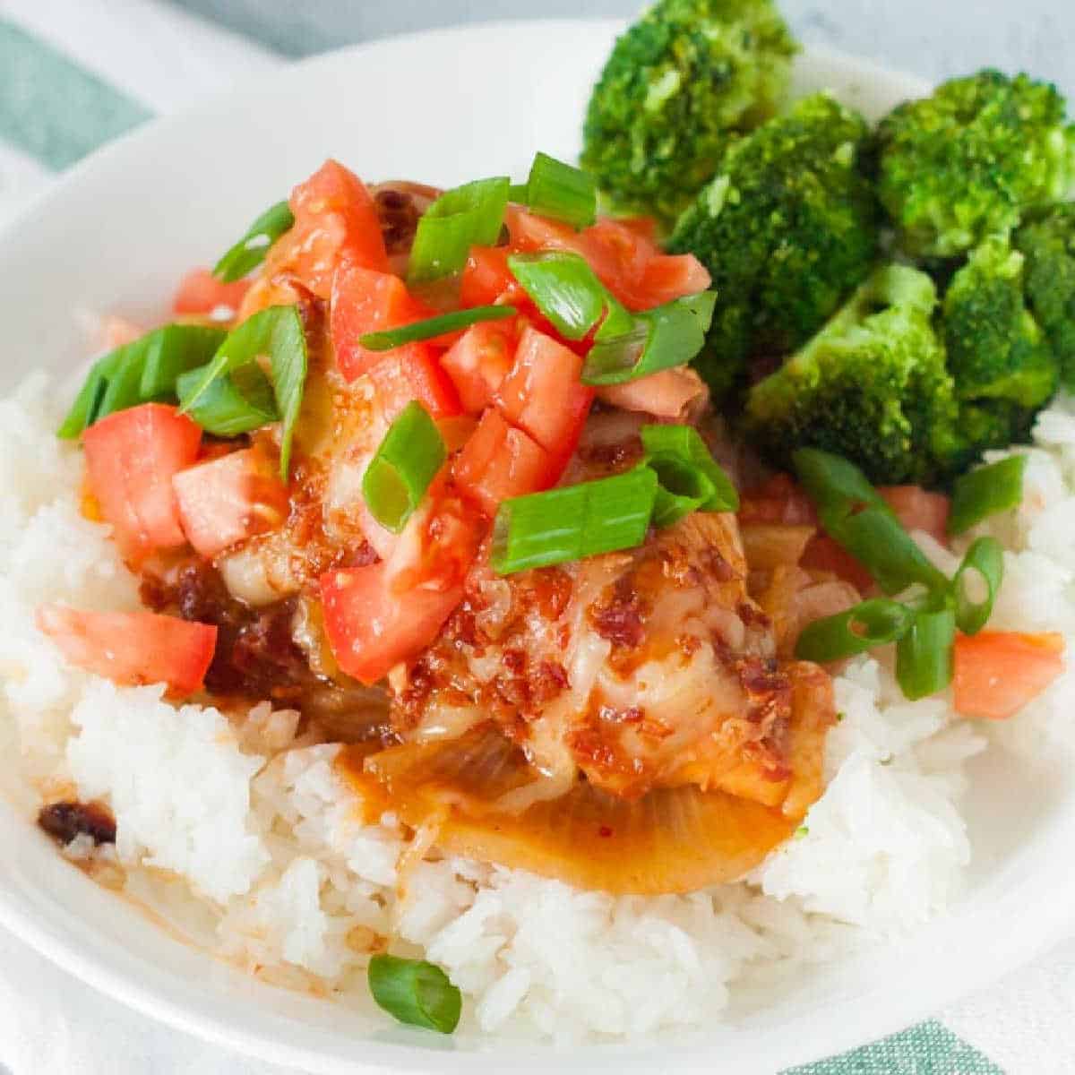 A white plate presents a delightful crock pot Monterey chicken recipe with steamed white rice, topped with juicy chicken drenched in red sauce and diced tomatoes. Sliced green onions add aroma, while broccoli florets nestle beside the savory dish.