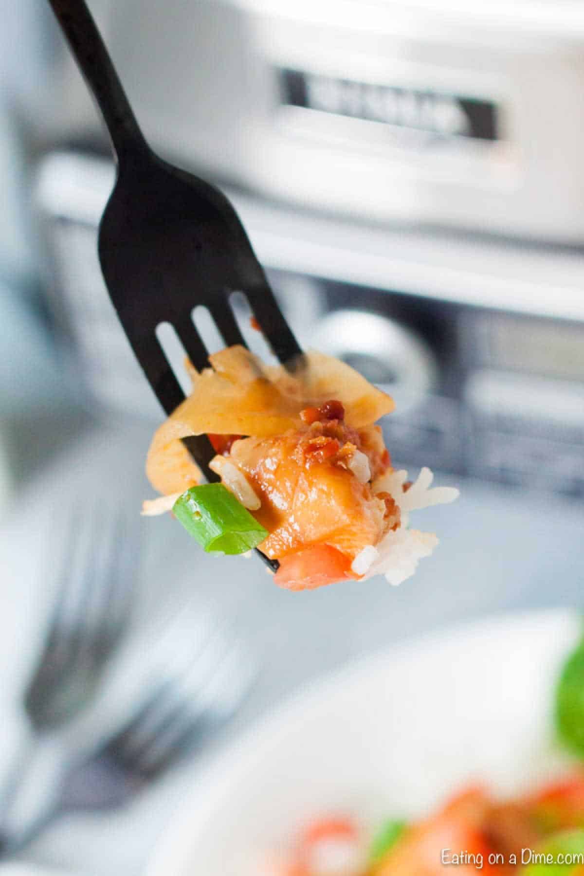 A close-up of a black fork holding a bite of Crock Pot Monterey Chicken, featuring rice, diced tomatoes, green bell pepper, and shredded chicken. A blurry plate and silver appliance are visible in the background.