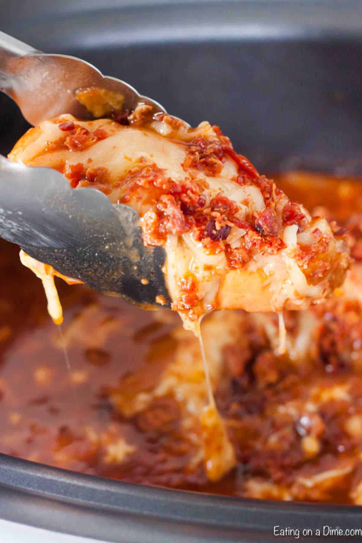 Tongs lift a portion of cheesy, saucy casserole from a dish, evoking the essence of a crock pot Monterey chicken recipe. Melted cheese and crispy bits top the bubbling delight, with rich red sauce gleaming in the background.